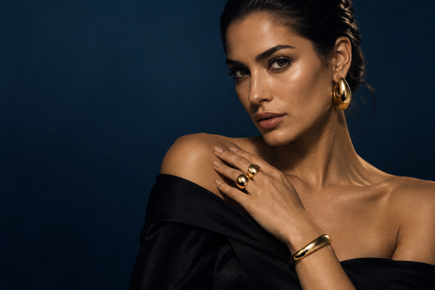 Woman wearing sculptural gold earrings, rings and a cuff against midnight navy